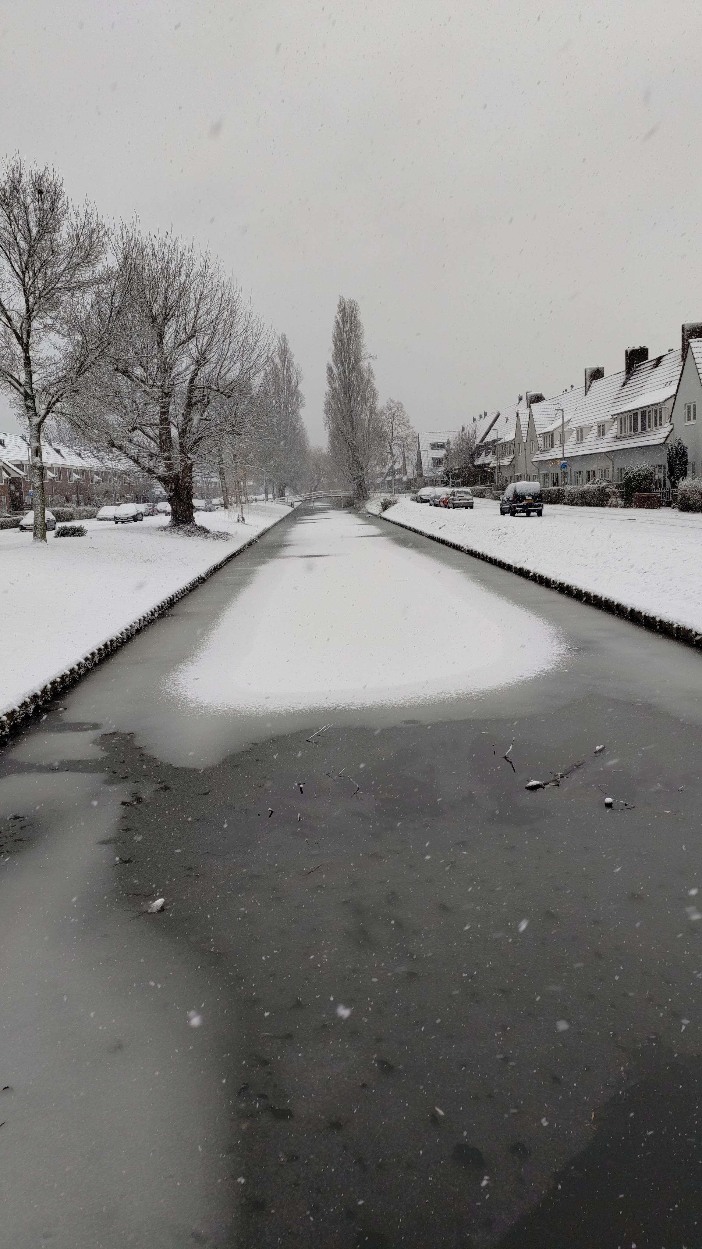 Photo of a partly frozen canal. The canal is in the middle, starting from the bottom of the picture it goes towards the top. On the sides there is the river bank, covered in snow, some naked trees and houses. The sky is grey.